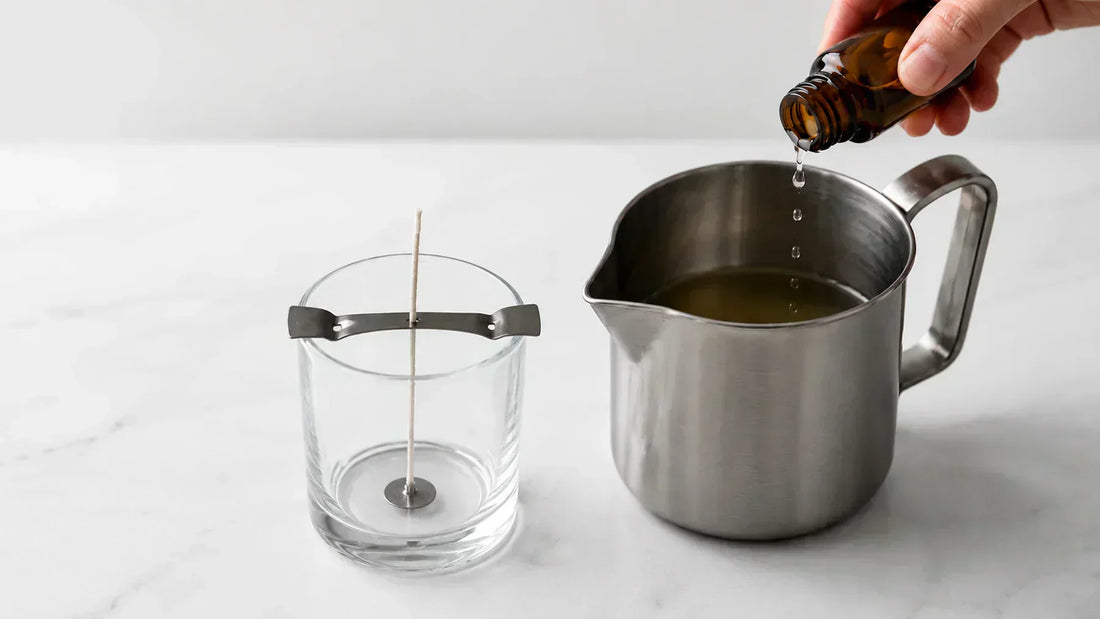 Fragrance oil being added to melted soy wax during handmade candle production with a glass candle jar and centered cotton wick