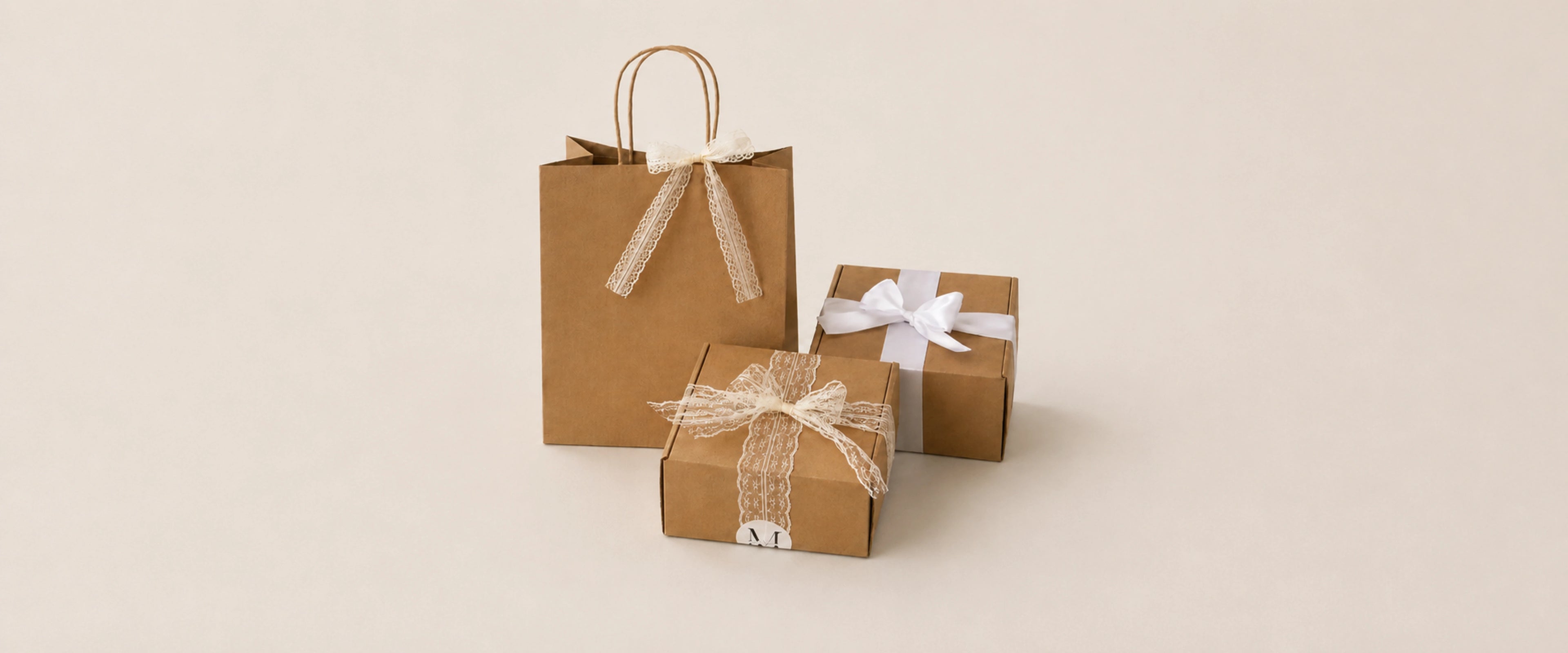 Three gift packaging items on a warm beige background: a large brown kraft paper shopping bag with twisted handles and a beige ribbon, a small brown gift box tied with a white ribbon and bow, and a medium brown gift box with a beige ribbon and bow.