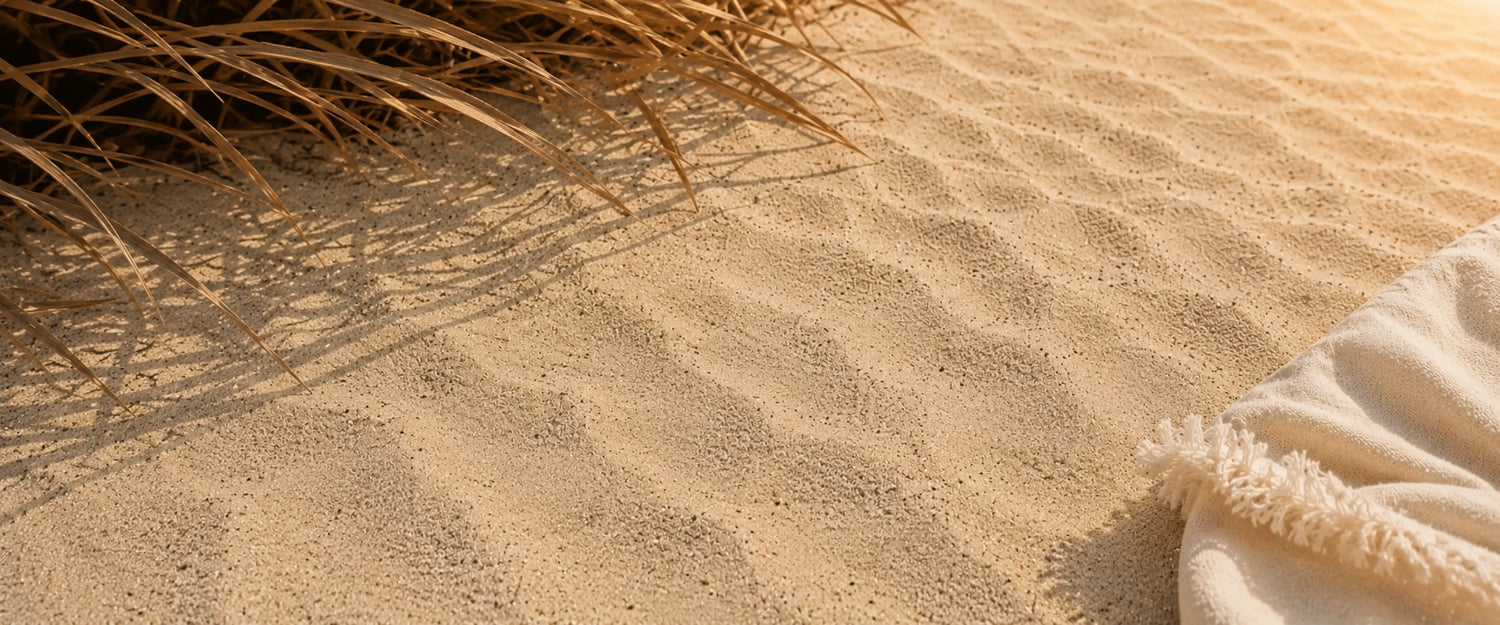 A close-up beach scene with rippled sand in the foreground, dry grass at the top left casting shadows, and a cream fringed textile corner at the bottom right.