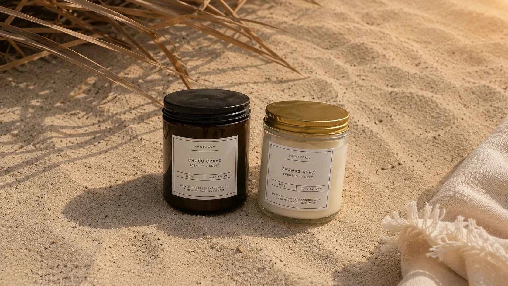Choco Crave scented soy candle in dark glass jar with black lid and Ananas Aura scented soy candle in clear glass jar with gold lid in warm beach setting